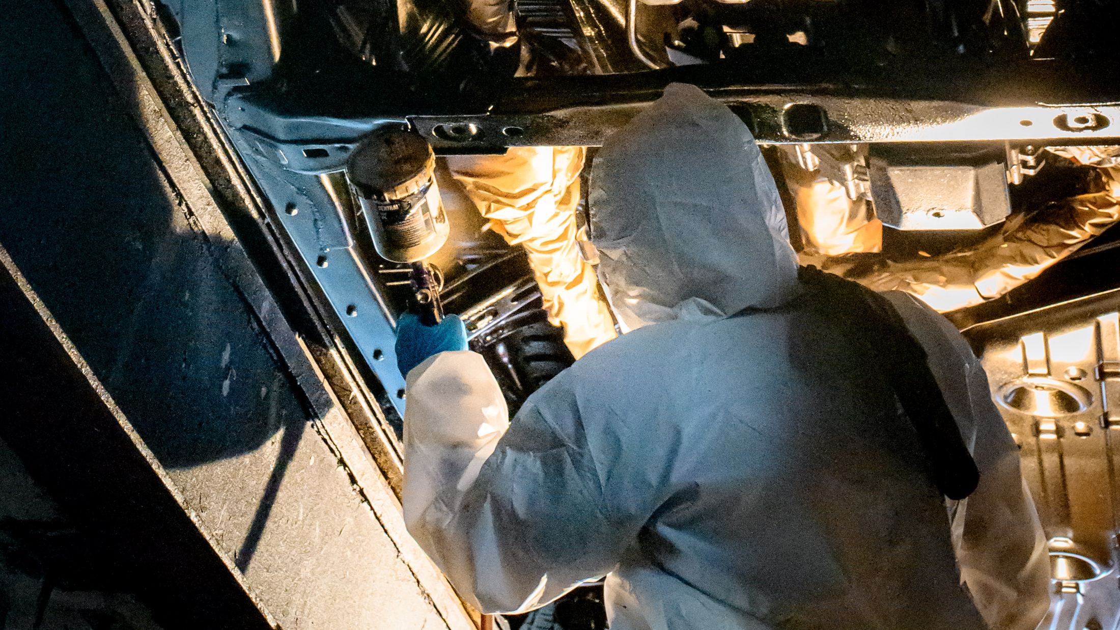 Technician in a protective suit spraying undercoating along the underside of a lifted vehicle